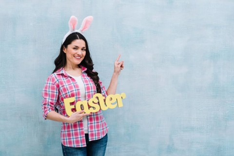 Portrait happy woman with bunny ears head holding easter word pointing finger upward blue background