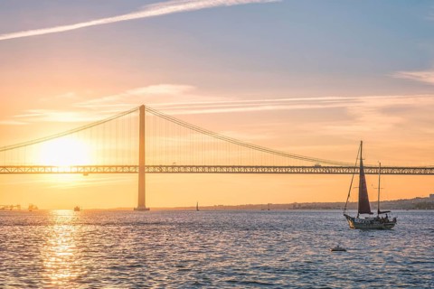 View of 25 de Abril Bridge famous tourist landmark of Lisbon connecting Lisboa and Almada over Tagus river with tourist yacht silhouette at sunset. Lisbon