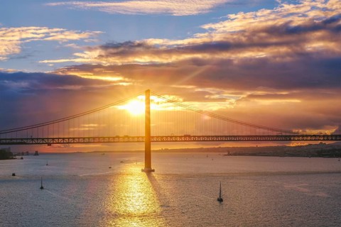 View of 25 de Abril Bridge famous tourist landmark of Lisbon connecting Lisboa and Almada on Setubal Peninsula over Tagus river with tourist yacht boats on sunset. Lisbon