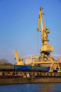 Colorful close vertical shot of river port crane loading open-top gondola cars on sunny day. Empty river drag boats or barges moored by pier. Empty cars ready for loading. Vertical image