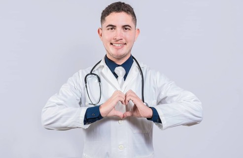 Friendly doctor making heart gesture with hands isolated. Young latin doctor making heart shape on isolated background
