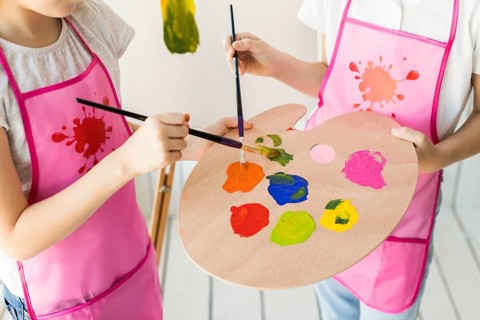 Elevated view two girls same pink apron mixing paint wooden palette with paint brush