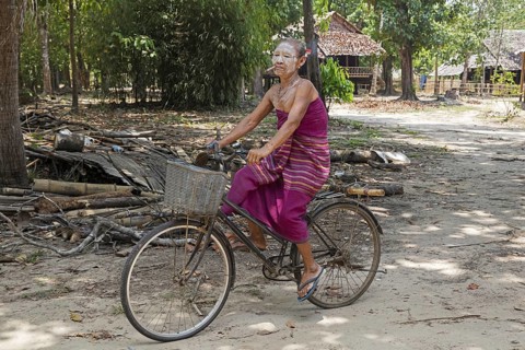 Old Burmese woman wearing thanaka and smoking a cigar while cycling in Kayin village near Hpa-an