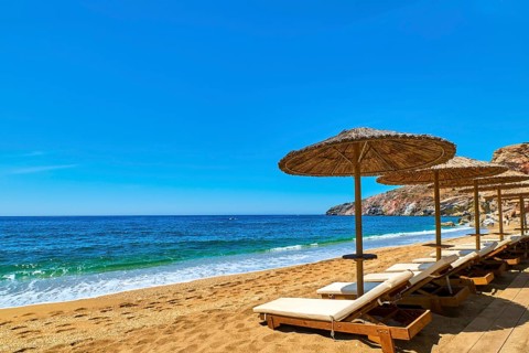 Beautiful empty beach with sun umbrellas and sunbeds. Perfect summer destination. Straw sunshades and sunbeds on the empty beach with blue sea and rocky hills. Vacations in Greece