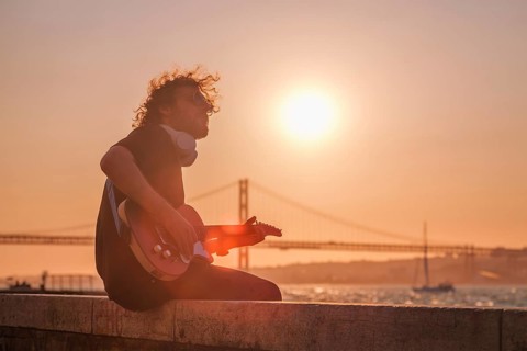 Hipster street musician in black playing electric guitar in the street on sunset on embankment with 25th of April bridge in background. Lisbon