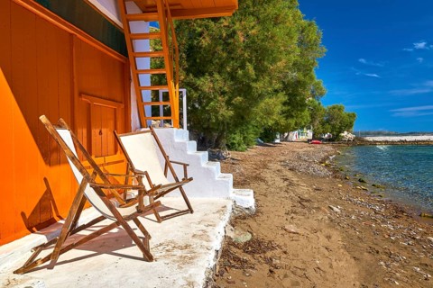 Two wooden beach chairs on porch of traditional Greek fisherman house just by sea tide. Whitewashed house walls and orange painted doors and stairs