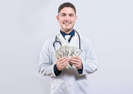 Latin young doctor holding money isolated. Cheerful doctor showing dollars isolated