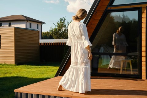 Playful woman in long white sundress looking at her reflection in the window
