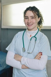 Smiling young female doctor medical nurse wearing white coat and stethoscope around neck standing in private clinic with arms crossed looking at camera. Vertical portrait