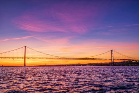 View of 25 de Abril Bridge famous tourist landmark of Lisbon connecting Lisboa and Almada over Tagus river with tourist yacht silhouette at sunset. Lisbon