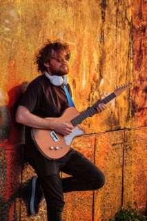 Hipster street musician in black playing electric guitar in the street on sunset leaning on a painted wall