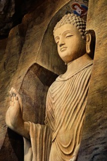 Statue of standing Buddha in monk clothes carved in limestone cave and lit by light. Right hand in adhay mudra gesture. Religion