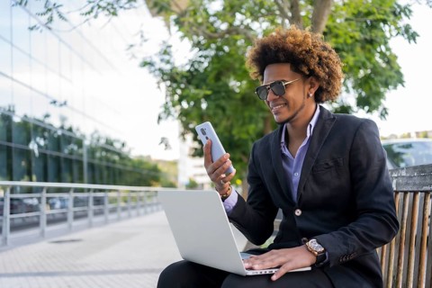 Cool businessman using mobile phone and laptop while working sitting on a urban park bench