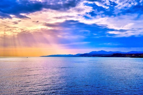 Colorful seascape with colorful clouds and bright sun rays cast over calm sea waters and hazy distant shores. Sunrise or sunset in Mediterranean sea