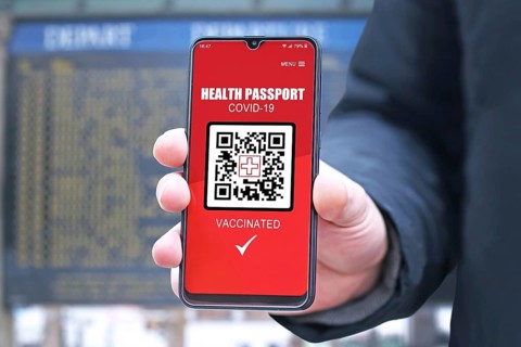 Corona virus vaccine passport on mobile phone device held by hand in front of air port or train station departure board to allow vaccinated people to travel