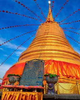 Stone board with with long message in Thai in front of big golden stupa or chedi decorated with festive red flags and textile in sunset sunlight in Wat Saket or Golden Mount temple