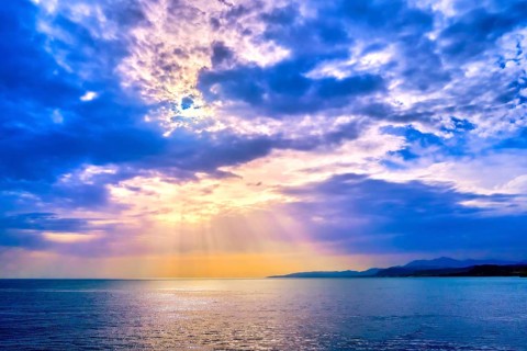 Beautiful seascape with colorful clouds and bright sun rays cast over calm sea waters and hazy distant shores. Sunrise or sunset in Mediterranean sea