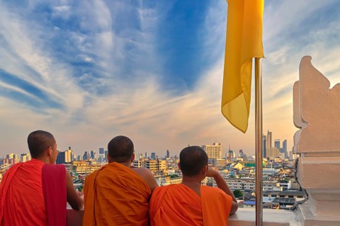 Three male Buddhist monks stand by white parapet wall of Wat Saket or Golden Mount temple and look at skyline of Bangkok