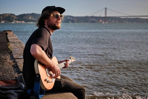 Hipster street musician in black playing electric guitar in the street sitting on pier embankment on sunset in Lisbon