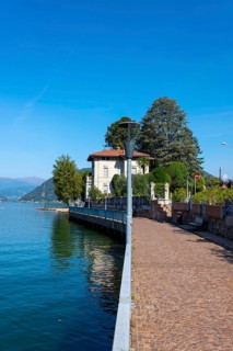 Walkway with Lamps and Railing and a House on the Waterfront on Lake Lugano with Mountain and Trees in a Sunny Summer Day in Porto Ceresio