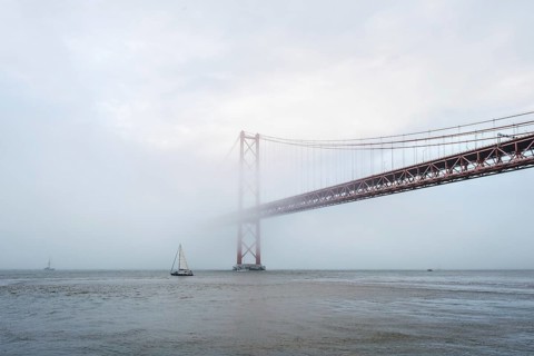 View of 25 de Abril Bridge famous tourist landmark of Lisbon connecting Lisboa and Almada in heavy fog mist wtih yacht boats passing under. Lisbon