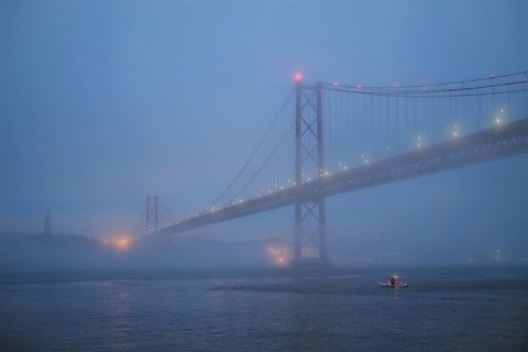 View of 25 de Abril Bridge famous tourist landmark of Lisbon connecting Lisboa and Almada in heavy fog mist wtih yacht boats passing under. Lisbon