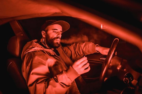 Rapper man in a cap in a car smoking at night in a garage lit with a red light in winter