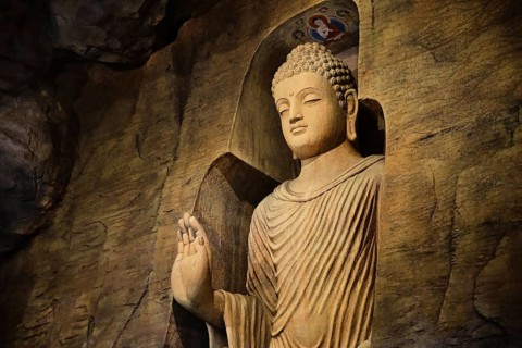 Statue of standing Buddha in monk clothes carved in limestone cave and lit by light. Right hand in adhay mudra gesture. Religion