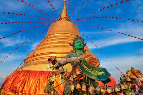 Statue of green-skinned Thai angel holding roll with message in Thai stand in front of big golden stupa in sunlight and at blue sky with clouds in Buddhist temple. Decorations with bells