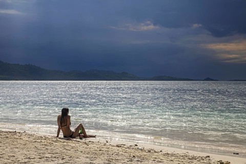 Female tourist in bikini sitting on tropical beach of the islet Gili Kedis and watching approaching thunderstorm over the island Lombok