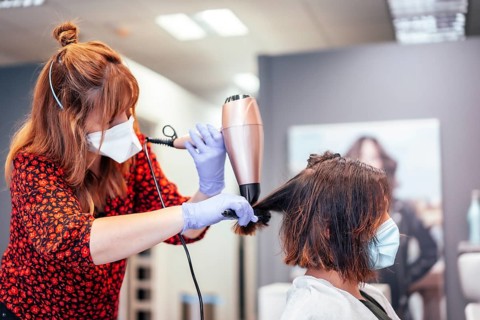 Blonde hairdresser with mask and gloves straightening client's brown hair with blow dryer. Reopening with security measures for hairdressers in the Covid-19 pandemic