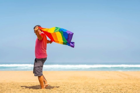 A gay person with a pink t-shirt and black cap with the LGBT flag on a beach