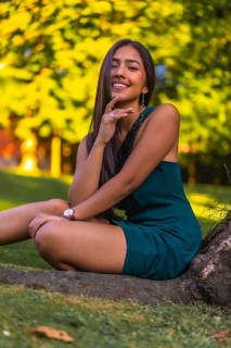 A young pretty brunette Latina with long straight hair leaning against a tree in a green dress. Portrait sitting on the grass next to a tree in the park