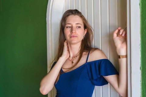 A young redhead Caucasian girl smiling in a blue dress next to a white door of a green house