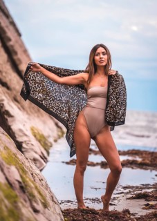 A dark-haired caucasian woman in a brown swimsuit and sarong on a natural background next to rocks and sea. In the summer sunset. Posing with the beautiful sarong next to the water