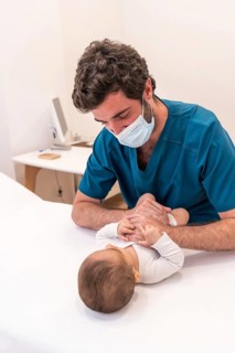 Frontal photo of a young doctor with facial mask examining a cute newborn baby in a clinic