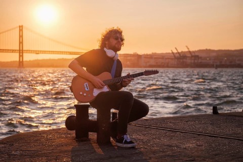 Hipster street musician in black playing electric guitar in the street on sunset on embankment with 25th of April bridge in background. Lisbon