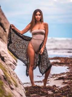 A dark-haired caucasian woman in a brown swimsuit and sarong on a natural background next to rocks and sea. In the summer sunset