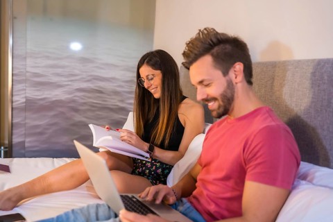 A handsome Caucasian couple lounging on the hotel bed and looking at a computer and a book. Summer lifestyle