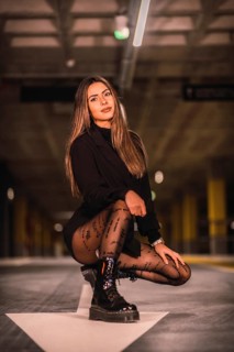 Caucasian brunette model with black jacket posing on the white arrow of the ground in an empty underground car park. Night urban session in the city