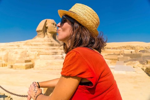 A young tourist in a red dress joking at the Great Sphinx of Giza and in the background the Pyramids of Giza