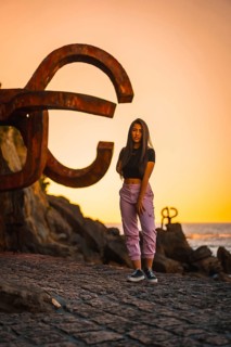 A young and pretty brunette Latina with long straight hair in a short black T-shirt and pink pants. Next to a sculpture of San Sebatian called Peine del Viento at sunset