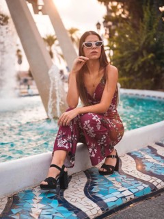 Portrait of a young brunette in a maroon floral dress and modern white sunglasses enjoying the summer in the golden hour in a city fountain