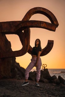 A young pretty brunette Latina with long straight hair in a short black T-shirt and pink pants. Perched next to a famous sculpture of Saint Sebatian called Peine del Viento