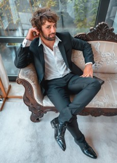 Street style with a young brunette caucasian man in a black suit and a white shirt in a luxurious hotel. Sitting on a traditional sofa looking at camera