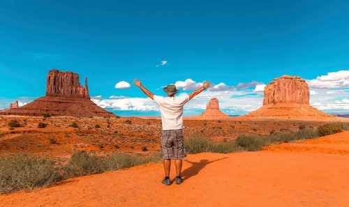 Man in white t-shirt and green hat with arms raised in Monument Valley National Park at The Mittens and Merrick Butte point. Utah