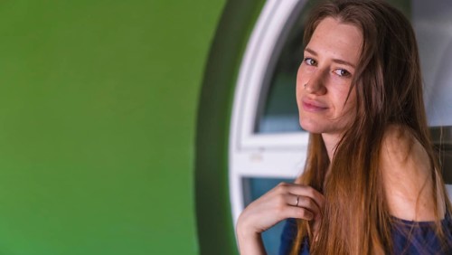 A smiling young pretty redhead Caucasian woman sitting in a blue dress next to a white sale of a green house
