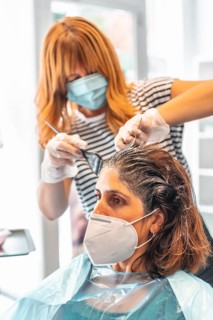 Blonde hairdresser with face mask giving the dark tint to the client at the hairdresser. Safety measures for hairdressers in the Covid-19 pandemic. New normal