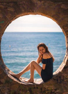 A young pretty red-haired Caucasian girl smiling sitting in a blue dress by the sea