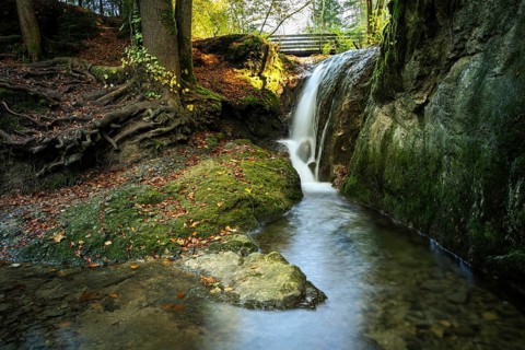 A small waterfall near the Gerats waterfall with moss-covered rocks and tree roots. The sun is shining in the background. Gerats